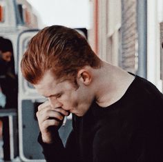 a man with red hair talking on a cell phone while standing next to a building