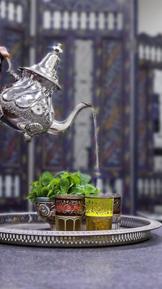 a silver teapot pouring water into small cups on a tray with basil and lemons