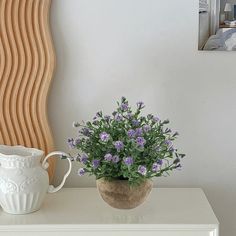 a white vase with purple flowers sitting on top of a table next to a wall