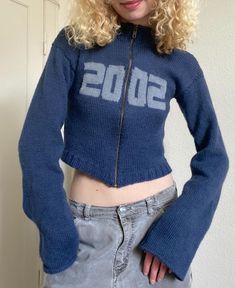 a woman with blonde hair wearing a blue sweater and grey jeans standing in front of a white wall