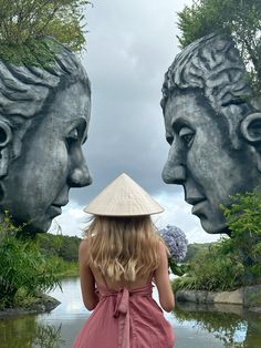 a woman wearing a hat looking at two statues in front of a body of water