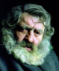 an old man with long grey hair and a beard wearing a black coat looking at the camera