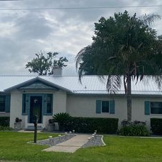 a white house with blue shutters and a palm tree in the front yard on a cloudy day