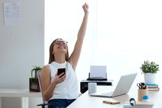 a woman sitting at a desk raising her arms in the air while holding a cell phone