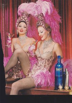 two women dressed in costumes sitting on a table next to bottles and glasses with pink feathers