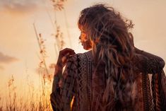 a woman standing in tall grass with her hair blowing in the wind and sunset behind her