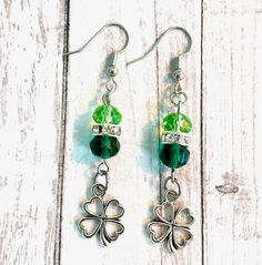 Feel the luck of the Irish this St. Patrick's Day with these beautiful, handmade shamrock earrings! These earrings include a metal alloy open shamrock or a green enamel filled shamrock paired with light and dark green crystal briolette beads and a rhinestone rondelle spacer bead. The open shamrock charm measures approximately 17mm by 12mm and the green enamel shamrock measures 19mm by 13mm. Dangle length of the earring is approximately 1.75-inches.Break out of the pandemic slump and get into the Green Drop Earrings With Dangling Charms, Dangle Earrings With May Birthstone In Metal, May Birthstone Dangle Earrings, Shamrock Earrings, Luck Of The Irish, Green Enamel, Green Crystals, Heart Jewelry, Spacer Beads