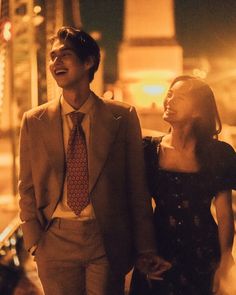 a man and woman are walking down the street at night, one is wearing a suit