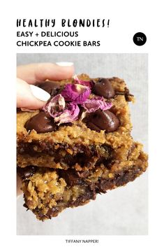 a hand holding two chocolate chip cookies with purple flowers on top, and one in the foreground