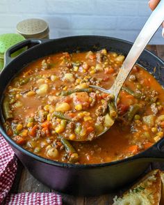 a spoon full of vegetable soup in a pot with bread on the side and a red checkered napkin next to it