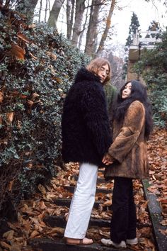 two women standing next to each other in front of some bushes and trees with leaves on the ground