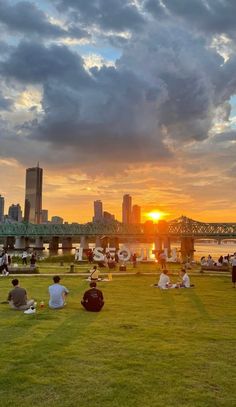 people are sitting on the grass in front of a bridge and some buildings at sunset