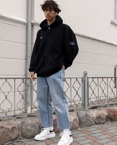 a young man standing on the sidewalk in front of a building wearing jeans and a hoodie