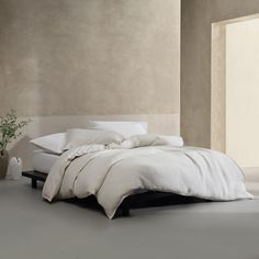 a bed with white sheets and pillows in a room next to a plant on the floor