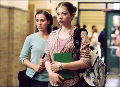 two young women are walking down the hallway with folders in their hands and backpacks on their backs