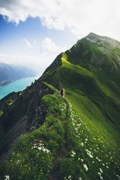 two people walking up the side of a mountain on a grassy hill with water in the background