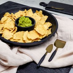 a black plate topped with guacamole and tortilla chips next to utensils