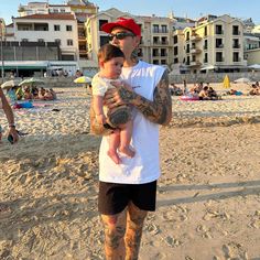 a man holding a baby on the beach in front of some buildings and people with their arms around each other