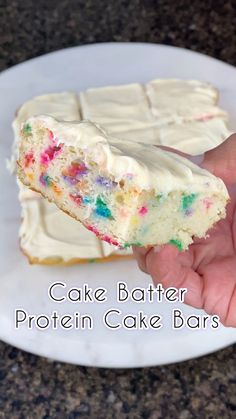 a hand holding a piece of cake batter protein cake bars on a white plate with rainbow sprinkles