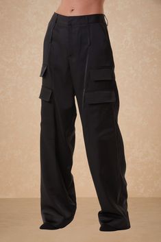 A person wearing Milan Satin Cargo Pants - Black from Noli Yoga, featuring multiple pockets, stands against a neutral background. The mid-rise cargo pants are loose-fitting and extend down to their feet. The upper torso is not visible in the image. Fall Staples, Cargo Pant, Sports Accessories, Satin Fabric, Black Pants, Best Sellers, Jumpsuit Dress, Milan, Mid Rise