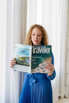 a woman in a blue dress is reading a travel magazine and looking at the camera