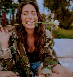 a woman in camo jacket sitting on bed holding up a flower to her mouth