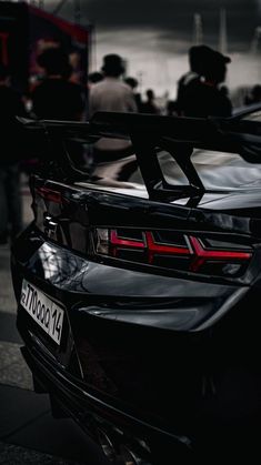 the back end of a black sports car with people standing around it in the background