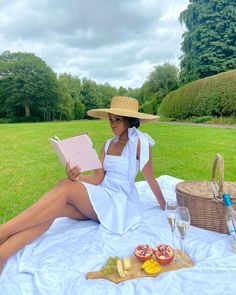 a woman in a white dress and straw hat sitting on a blanket reading a book