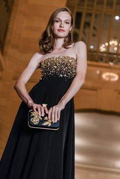 a woman in a black evening gown holding a gold clutch on her hand and looking off to the side