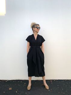 a woman standing in front of a white wall wearing a black dress with a bow at the waist