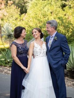 two women and a man standing next to each other in front of trees with bushes