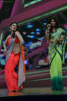 two women in sari on stage with one holding a microphone and the other singing