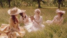 three women in dresses and hats sitting on the grass reading books while one looks at her cell phone