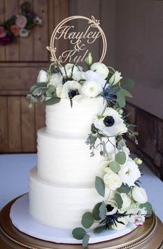 a wedding cake with white flowers and greenery on top is sitting on a table