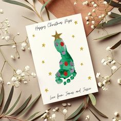 a card with a christmas tree on it sitting next to white flowers and greenery