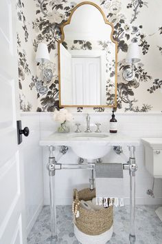 a white bathroom with floral wallpaper and mirror above the sink, along with a wicker basket on the floor