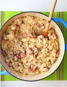 a blue pot filled with rice and meat on top of a colorful place mat next to a wooden spoon