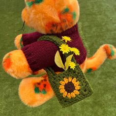 a teddy bear wearing a sweater and holding a crocheted bag with flowers on it