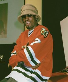 a man in an orange hockey jersey and hat smiles while sitting on a red bench