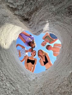 four women in bikinis are standing in a heart shaped hole on the beach, looking up at the camera