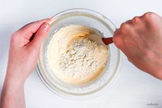 two hands mixing ingredients in a glass bowl on top of a white countertop with a wooden spoon