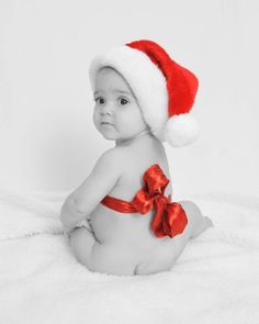 a baby wearing a santa hat and sitting on a white blanket with a red ribbon