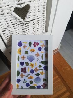 a person holding up a white frame with colorful leaves on it in front of a wicker basket