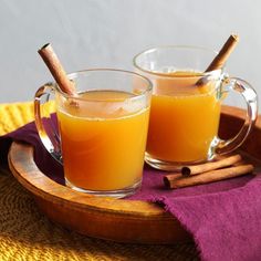 two mugs filled with orange juice and cinnamon sticks on a wooden tray next to a purple towel