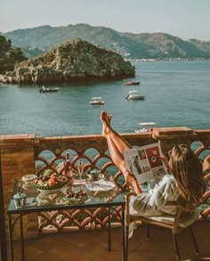 a woman sitting at a table reading a magazine overlooking the ocean and boats in the water