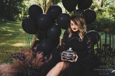 a woman in a black dress sitting on a chair holding a purse and balloons behind her