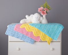 a crocheted blanket and teddy bear sitting on a dresser with flowers in a vase