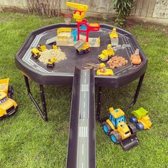 a toy table with construction vehicles on it in the grass next to a small road