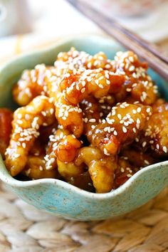 a blue bowl filled with chicken and sesame seeds next to chopsticks on a table