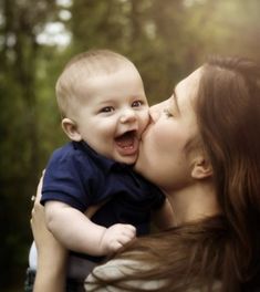 a woman holding a baby in her arms and kissing it's face with trees in the background